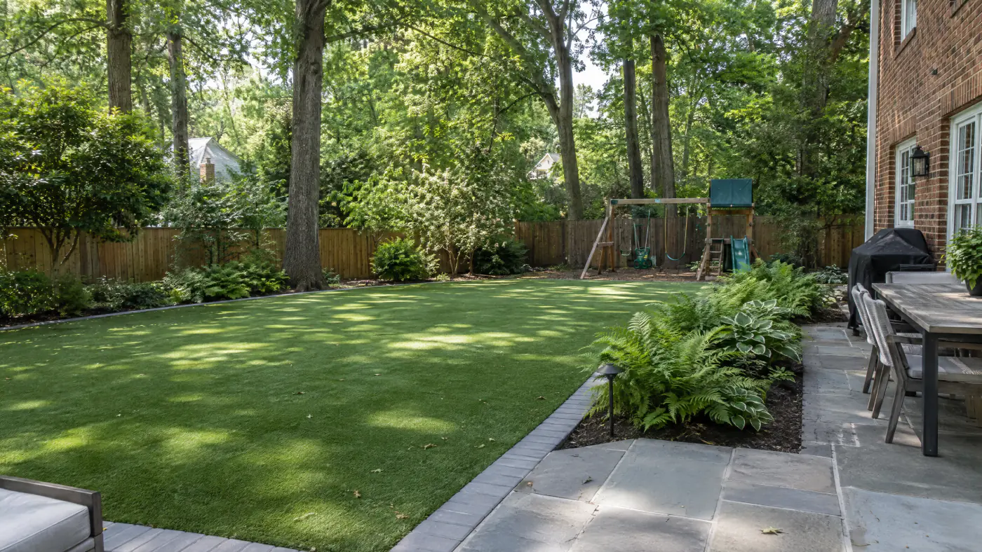 Artificial turf lawn with a paver border in a shaded backyard beneath mature oaks behind a brick colonial
