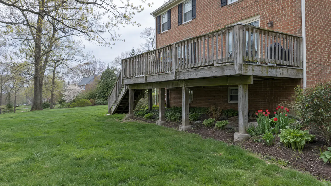 Weathered gray pressure-treated deck behind a brick colonial home in early April with tulips blooming below it