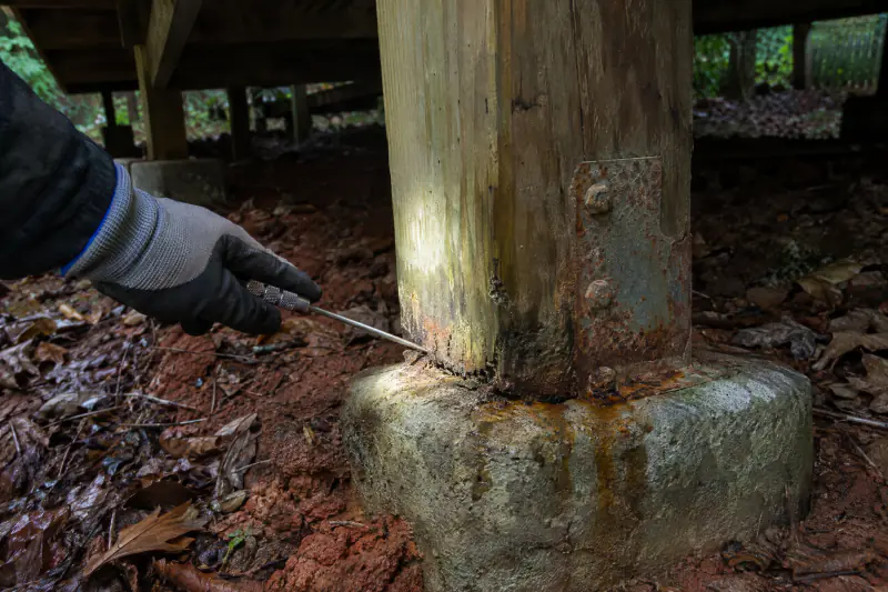 Awl probing a deck post base at a concrete footing with rust-stained hardware during a spring deck inspection