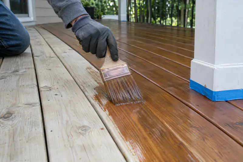 Brush applying semi-transparent stain to clean deck boards, with stained and bare boards side by side