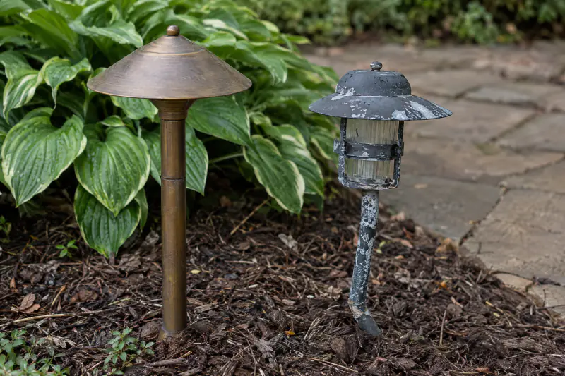 Solid brass path light with weathered patina beside a corroded aluminum fixture on a mulched bed edge
