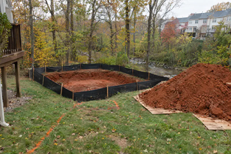 Silt fence staked around a patio excavation on a sloped Northern Virginia backyard with woods and a stream below