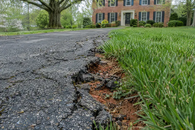 Cracked and rutted asphalt driveway edge crumbling into a tall fescue lawn beside a brick home in Northern Virginia