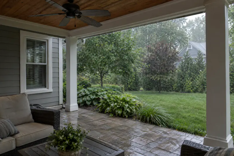 Open covered porch with a stained wood ceiling and fan over a paver patio during a summer rain shower