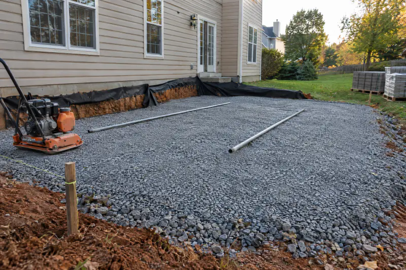 Compacted crusher-run paver base with geotextile at the edges, steel screed pipes and a plate compactor before bedding sand