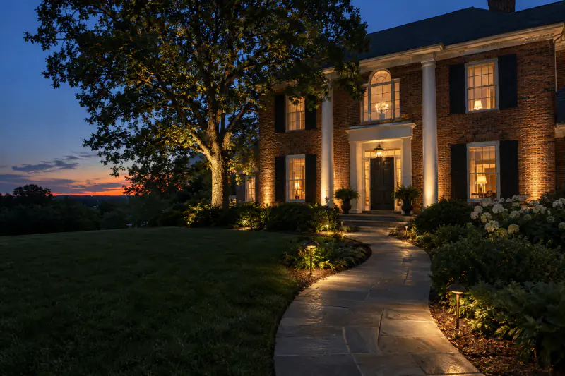 Brick colonial home at dusk with landscape lighting on walkway, facade and oak tree