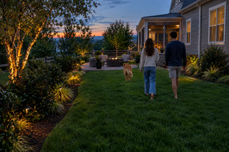 Couple and dog walking across a lawn and landscape with lighting at dusk