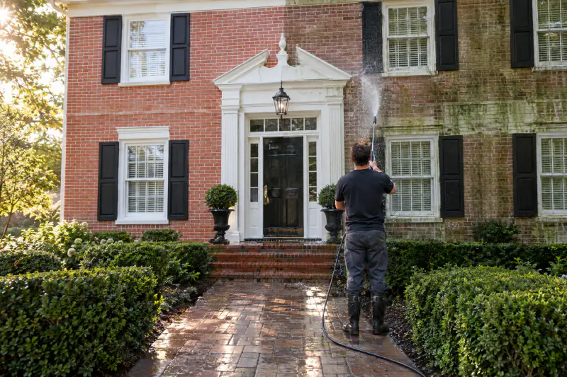 Technician soft-washing a brick colonial home, the cleaned half gleaming beside the algae-streaked half